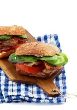 Delicious Ciabatta Sandwiches with Bacon, Tomato, Lettuce and Sauces on Cutting Board and Checkered Napkin isolated on white background