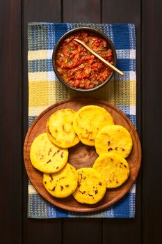 Overhead shot of arepas on wooden plate with Colombian hogao sauce (tomato and onion cooked) in bowl. Arepas are made of yellow or white corn meal and are traditionally eaten in Colombia and Venezuela. Photographed on dark wood with natural light.