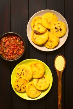 Overhead shot of arepas on plates with Colombian hogao sauce (tomato and onion cooked) in bowl. Arepas are made of yellow or white corn meal and are traditionally eaten in Colombia and Venezuela. Photographed on dark wood with natural light.