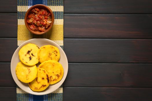 Overhead shot of arepas on plate with Colombian hogao sauce (tomato and onion cooked) in bowl. Arepas are made of yellow or white corn meal and are traditionally eaten in Colombia and Venezuela. Photographed on dark wood with natural light.