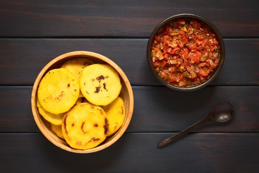 Overhead shot of arepas in wooden bowl with Colombian hogao sauce (tomato and onion cooked) and a spoon. Arepas are made of yellow or white corn meal and are traditionally eaten in Colombia and Venezuela. Photographed on dark wood with natural light.