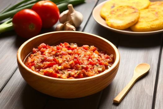 Colombian hogao or criollo sauce (salsa criolla) made of cooked onion and tomato, served as accompaniment to traditional dishes, with ingredients and arepas in the back, photographed on dark wood with natural light (Selective Focus, Focus in the middle of the sauce)