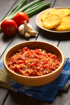 Colombian hogao or criollo sauce (salsa criolla) made of cooked onion and tomato, served as accompaniment to traditional dishes, with ingredients and arepas in the back, photographed on dark wood with natural light (Selective Focus, Focus in the middle of the sauce)