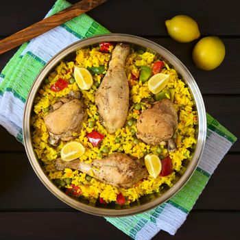 Overhead shot of a pot of chicken paella, a traditional Valencian (Spanish) rice dish made of rice, chicken, peas and capsicum and served with lemon, photographed on dark wood with natural light