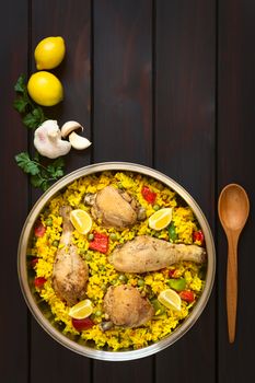 Overhead shot of a pot of chicken paella, a traditional Valencian (Spanish) rice dish made of rice, chicken, peas, capsicum and served with lemon, photographed on dark wood with natural light
