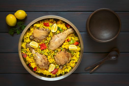 Overhead shot of a pot of chicken paella, a traditional Valencian (Spanish) rice dish made of rice, chicken, peas and capsicum and served with lemon, photographed on dark wood with natural light