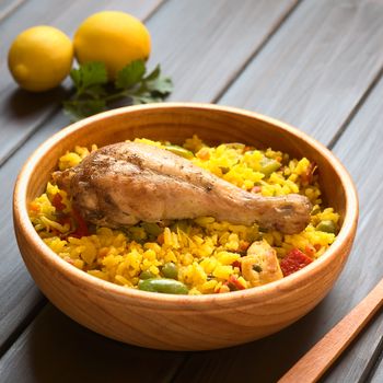 Wooden bowl of chicken paella, a traditional Valencian (Spanish) rice dish made of rice, chicken, peas and capsicum , photographed on dark wood with natural light (Selective Focus, Focus on the chicken thigh)