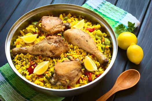 A pot of chicken paella, a traditional Valencian (Spanish) rice dish made of rice, chicken, peas, capsicum and served with lemon, photographed on dark wood with natural light (Selective Focus, Focus on the middle of the dish)