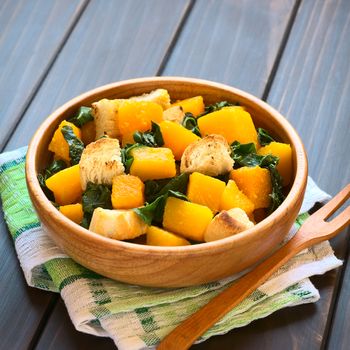 Pumpkin and chard salad with croutons served in wooden bowl, photographed on dark wood with natural light (Selective Focus, Focus in the middle of the salad) 