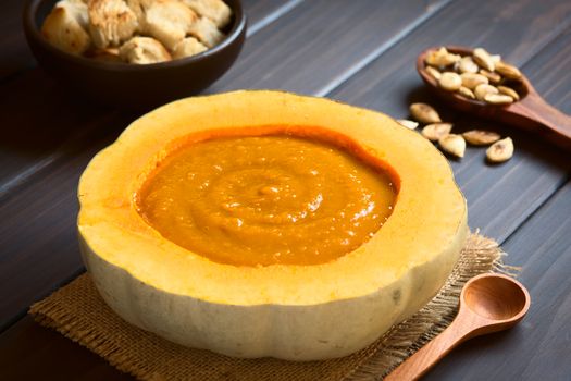 Cream of pumpkin soup served in half a pumpkin, photographed on dark wood with natural light (Selective Focus, Focus on the middle of the soup) 