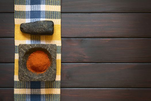 Paprika powder spice in stone mortar with pestle on kitchen towel, photographed overhead on dark wood with natural light
