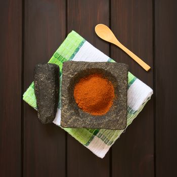Paprika powder spice in stone mortar with pestle on kitchen towel, photographed overhead on dark wood with natural light