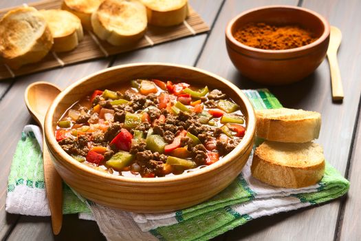 Vegan goulash made of soy meat (textured vegetable protein), capsicum, tomato and onion, served in wooden bowl accompanied by toasted bread, photographed with natural light (Selective Focus, Focus one third into the dish)