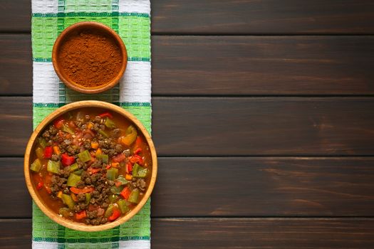 Vegan goulash made of soy meat (textured vegetable protein), capsicum, tomato and onion in wooden bowl, paprika powder in small bowl, photographed overhead on dark wood with natural light