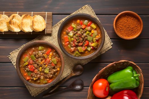 Vegan goulash made of soy meat (textured vegetable protein), capsicum, tomato and onion served in rustic bowls, toasted bread, paprika powder, fresh vegetables on the side, photographed overhead on dark wood with natural light