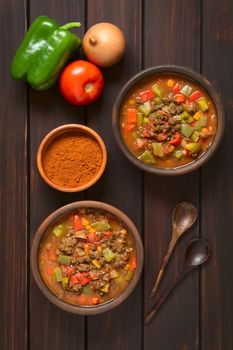 Vegan goulash made of soy meat (textured vegetable protein), capsicum, tomato and onion served in rustic bowls, paprika powder, fresh vegetables, spoons on the side, photographed overhead on dark wood with natural light