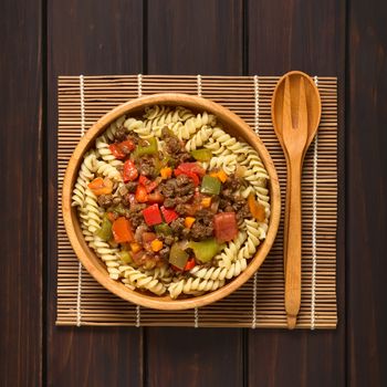 Vegan goulash made of soy meat (textured vegetable protein), capsicum, tomato and onion served on fusilli pasta in wooden bowl, spoon and fork on the side, photographed overhead on dark wood with natural light