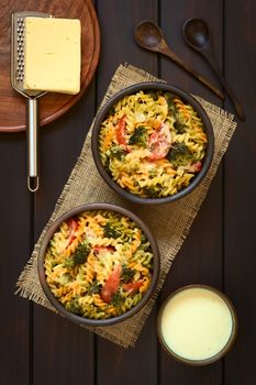 Baked tricolor fusilli pasta and vegetable (broccoli, tomato) casserole in rustic bowls, cream sauce, spoons, grater and cheese on the side, photographed overhead on dark wood with natural light