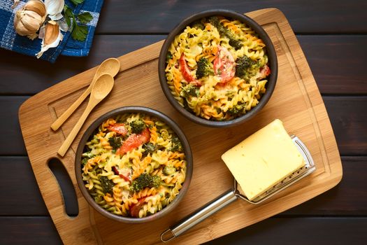 Baked tricolor fusilli pasta and vegetable (broccoli, tomato) casserole in rustic bowls, with grater, cheese and spoons on wooden board, photographed overhead on dark wood with natural light