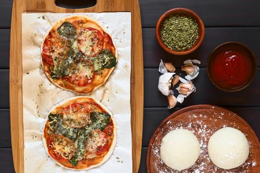 Homemade spinach and tomato pizza on baking paper on wooden board, pizza dough, dried oregano, tomato sauce, garlic on the side, photographed overhead on dark wood with natural light