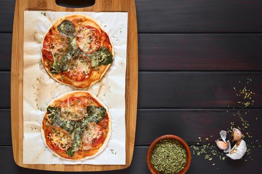 Homemade spinach and tomato pizza on baking paper on wooden board, dried oregano and garlic on the side, photographed overhead on dark wood with natural light