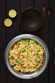 Vegetarian couscous salad made with bell pepper, tomato, cucumber, red onion and sweet corn kernels, served in salad bowl with rustic bowls, wooden spoons and lemon on the side. Photographed overhead on dark wood with natural light.