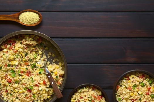 Vegetarian couscous salad made with bell pepper, tomato, cucumber, red onion and sweet corn kernels, raw couscous on wooden spoon on the top. Photographed overhead on dark wood with natural light.