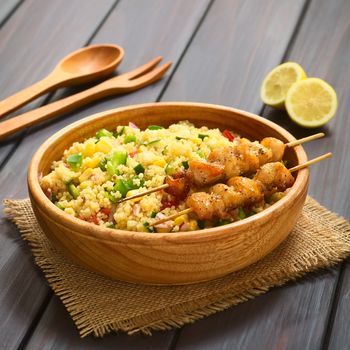 Couscous salad made with bell pepper, tomato, cucumber, red onion and sweet corn kernels, served with baked chicken on skewer in wooden bowl. Photographed on dark wood with natural light. (Selective Focus, Focus one third into the dish)