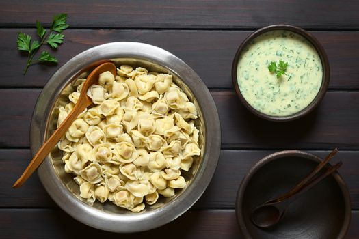 Cooked tortellini stuffed with cheese in bowl with parsley cream sauce and rustic bowls to serve, photographed overhead on dark wood with natural light