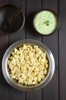 Cooked tortellini stuffed with cheese in bowl with parsley cream sauce and rustic bowls to serve, photographed overhead on dark wood with natural light