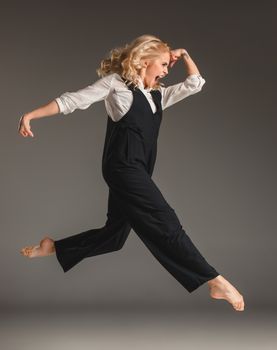 Beauty blond woman in a black suit and white shirt in ballet jump on a gray background