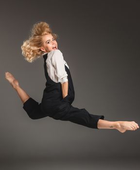 Beauty blond woman in a black suit and white shirt in ballet jump on a gray background