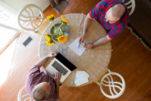 Discussion between two men seated at a round table decorated with yellow sunflowers using a laptop computer and paper documents, overhead view