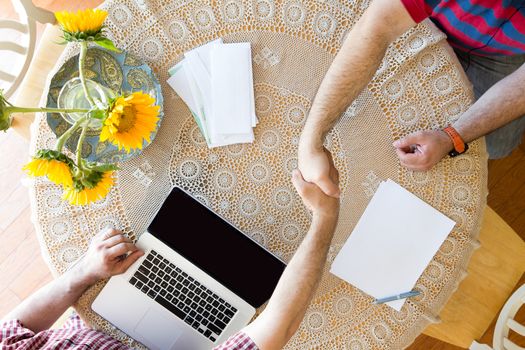 Overhead view of two men sitting at a table decorated with colorful yellow sunflowers shaking hands on an agreement, view of their hands and a laptop