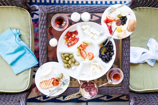 Unfinished Turkish breakfast on a patio table with a serving of fried eggs with a selection of fresh tomato, olives, cheese and mugs of Turkish tea, overhead view with napkins on chairs