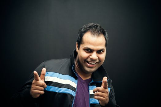 Portrait of young happy Indian man with positive gesture pointing at camera over dark background