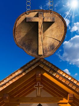 Wooden Christian cross on a section of tree trunk, hanging from a metal chain. In the background wooden roof of a mountain church
