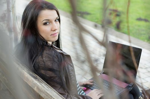 Young beautiful brunette student in profile sits on bench about university and works behind laptop.