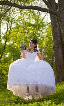 Portrait of a beautiful bride in white wedding dress sitting on swing outdoors