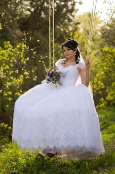 Portrait of a beautiful bride in white wedding dress sitting on swing outdoors