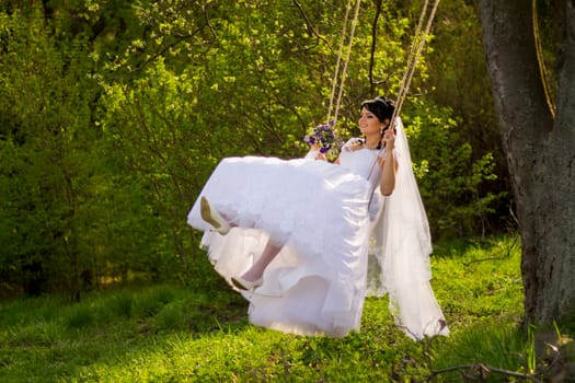 Portrait of a beautiful bride in white wedding dress sitting on swing outdoors