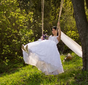 Portrait of a beautiful bride in white wedding dress sitting on swing outdoors