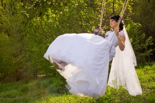 Portrait of a beautiful bride in white wedding dress sitting on swing outdoors