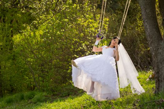 Portrait of a beautiful bride in white wedding dress sitting on swing outdoors