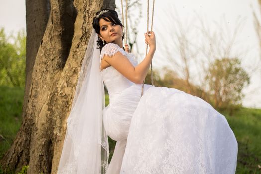 Portrait of a beautiful bride in white wedding dress sitting on swing outdoors