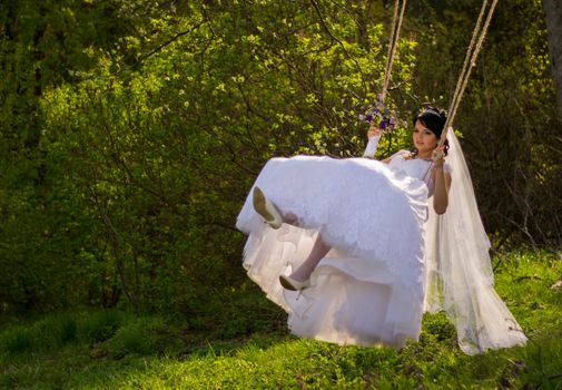 Portrait of a beautiful bride in white wedding dress sitting on swing outdoors