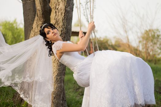 Portrait of a beautiful bride in white wedding dress sitting on swing outdoors