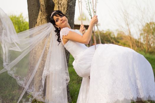 Portrait of a beautiful bride in white wedding dress sitting on swing outdoors