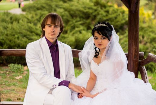 Couple in love bride and groom posing sitting on wooden bench in gazebo in their wedding day in summer.