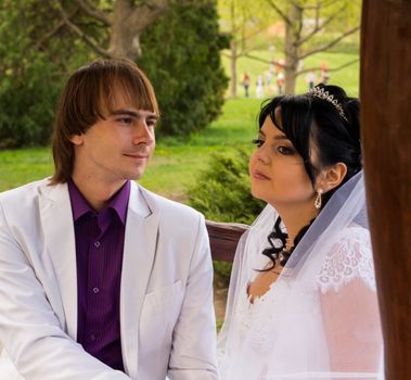 Couple in love bride and groom posing sitting on wooden bench in gazebo in their wedding day in summer.